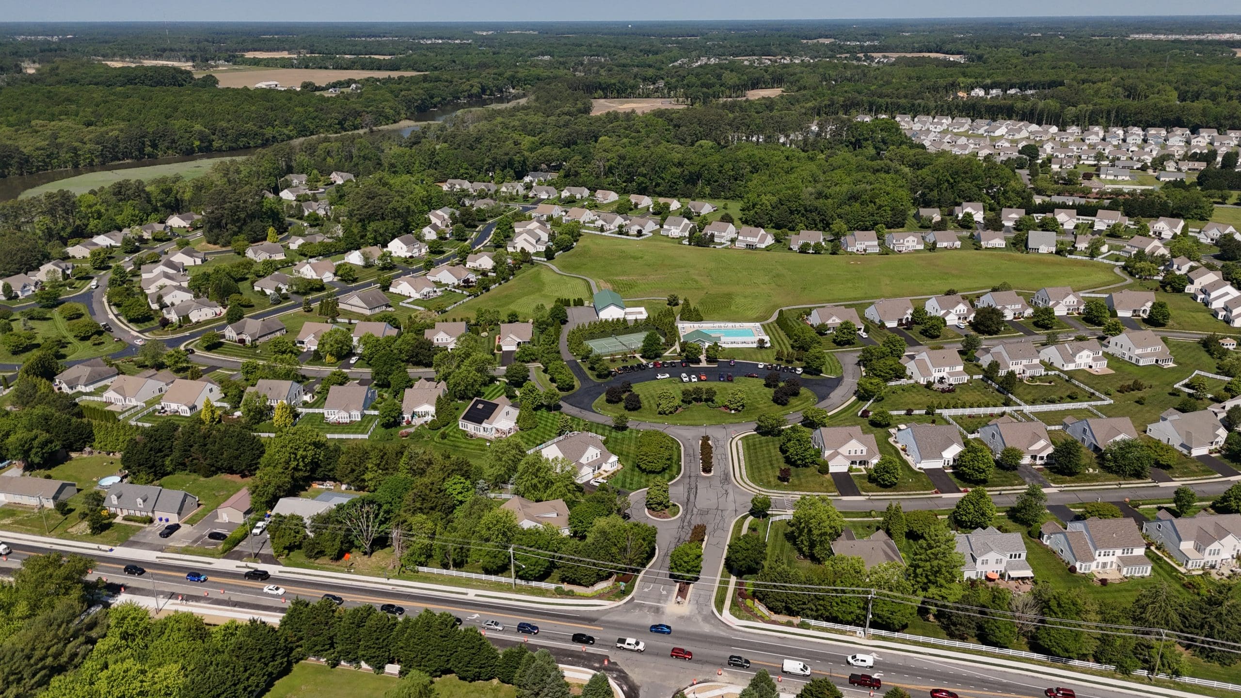 Aerial view of a suburban neighborhood with curved streets, rows of houses, green yards, and a central park area with a pool and tennis courts.