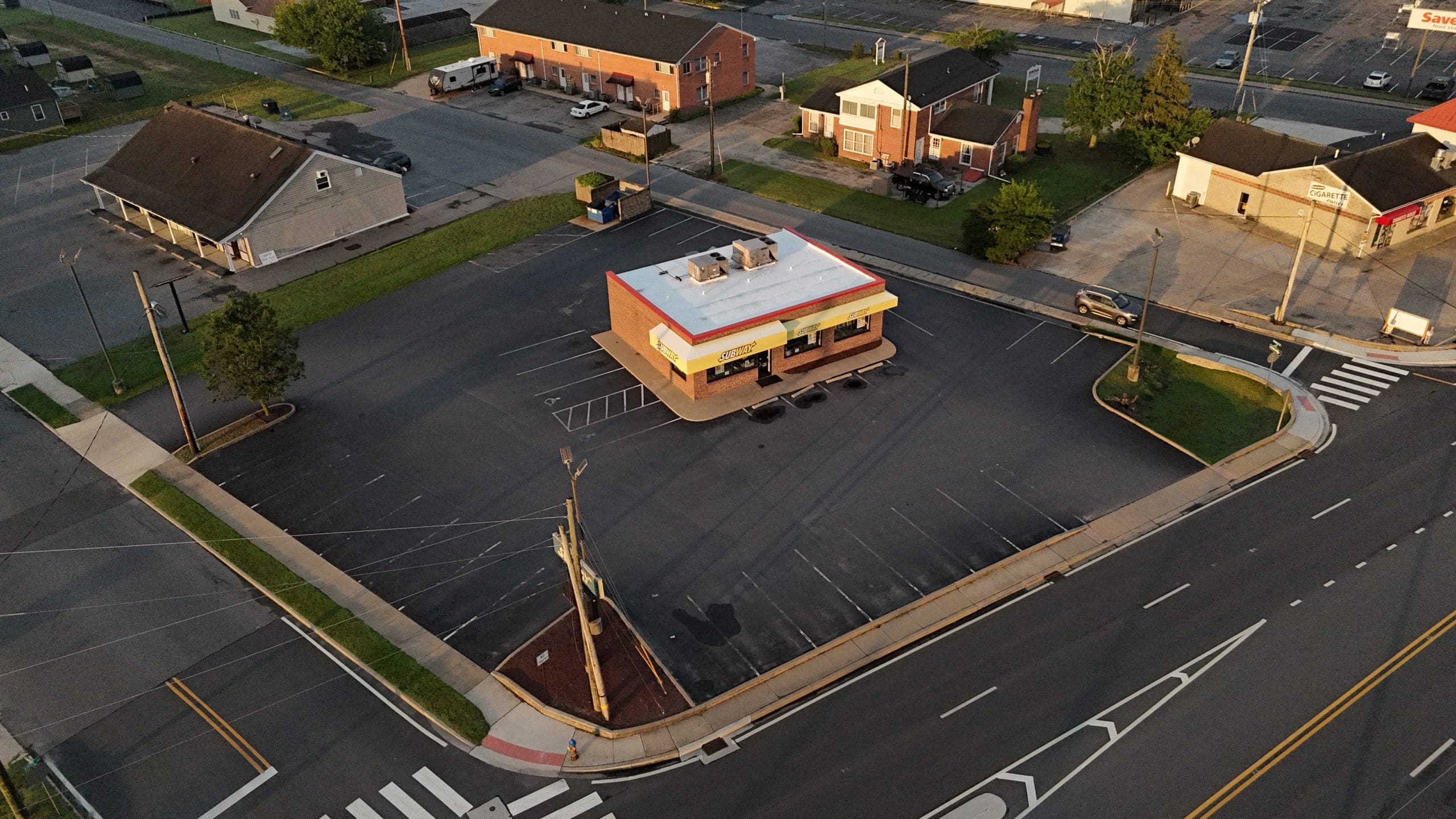 Aerial view of a brick Subway restaurant in a large empty parking lot, surrounded by roads and small houses.