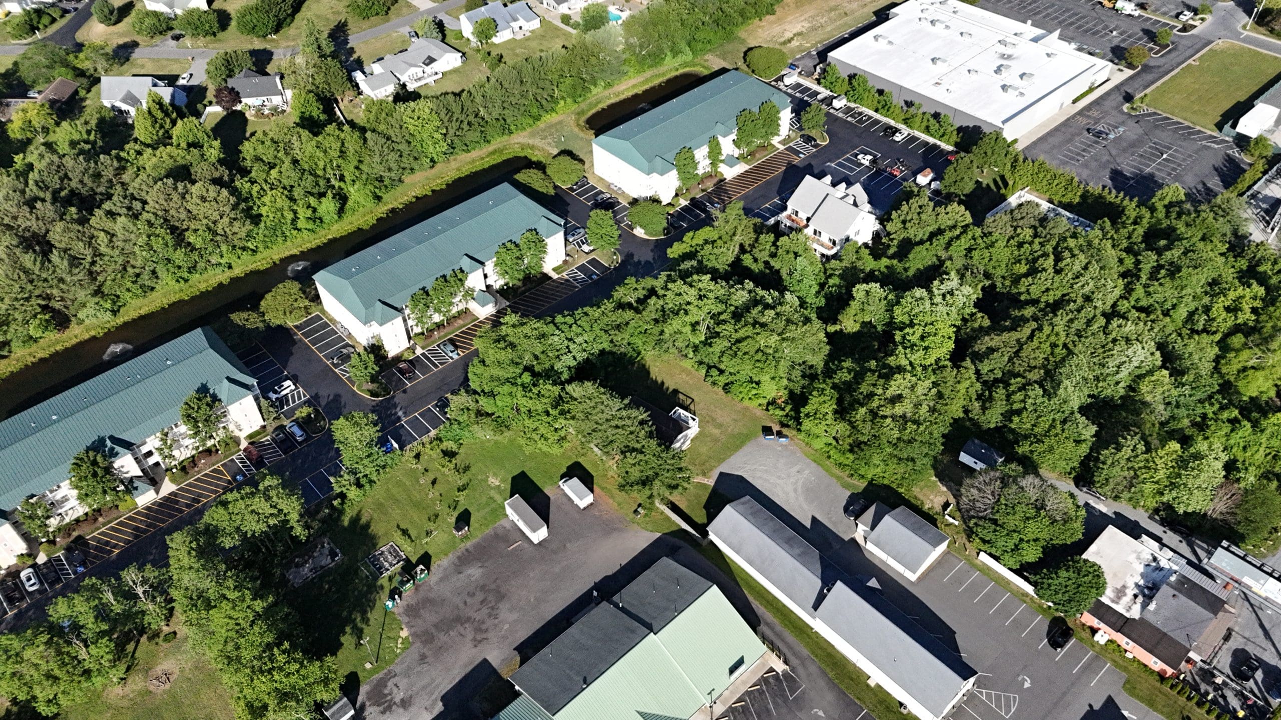 Aerial view of a green-roofed apartment complex with a canal beside it and surrounding trees and parking lots.